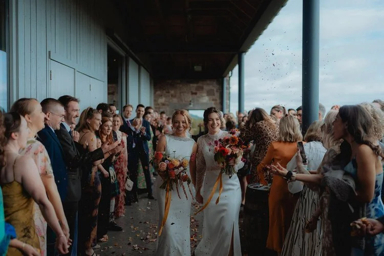 Two brides stepping into a storm of cheers and colour, captured with F.D Young Photography’s trademark blend of atmosphere and emotion. Their smiles say everything—joy that spills outwards, reaching every guest lining the aisle. With flowers in hand 