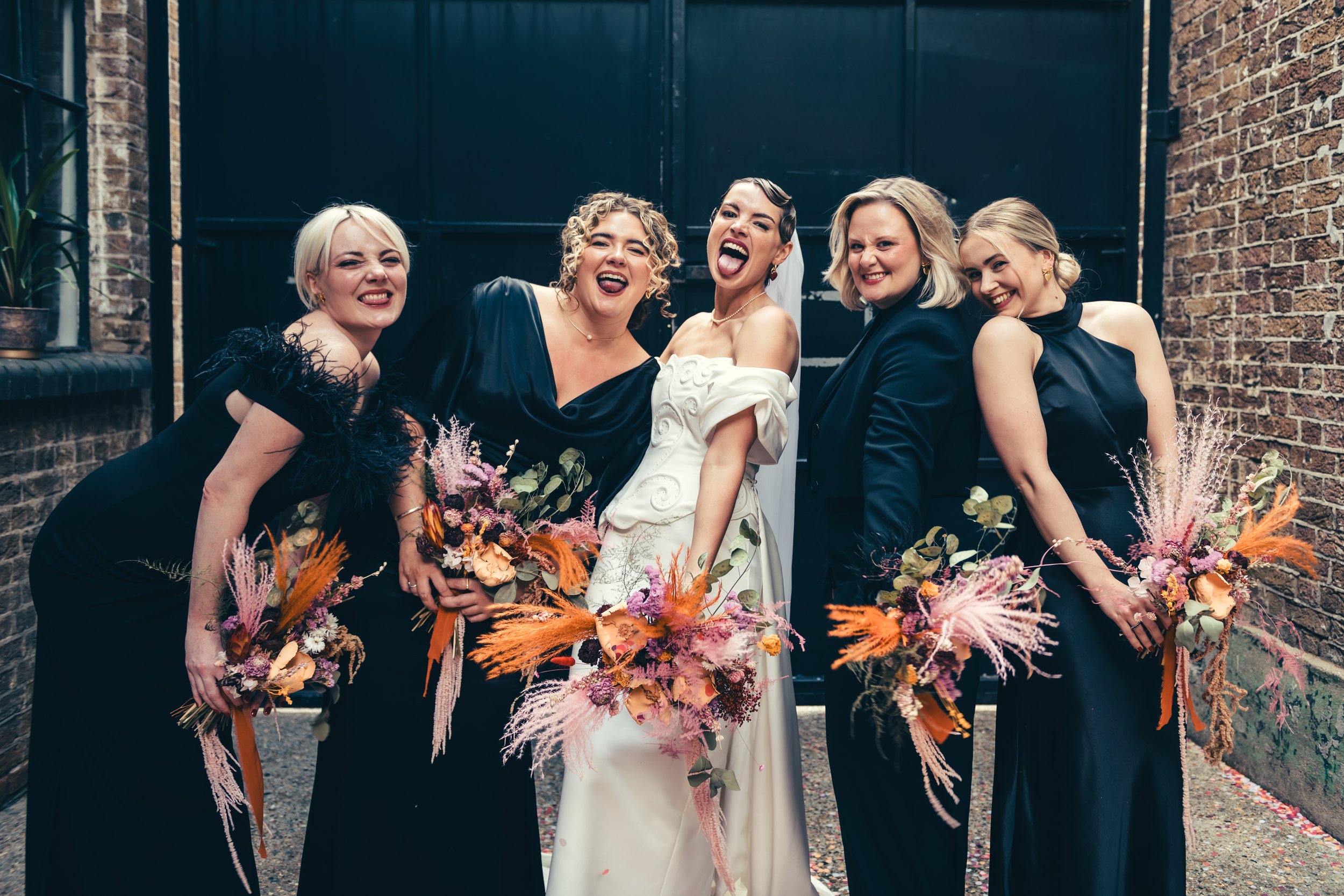 A joyful bridal party captured with makeup by Powder Blue Artistry, featuring glowing skin, bold lips, and elegant natural tones. The cohesive beauty styling enhances each look, complementing the modern black bridesmaid dresses and vibrant bouquets f