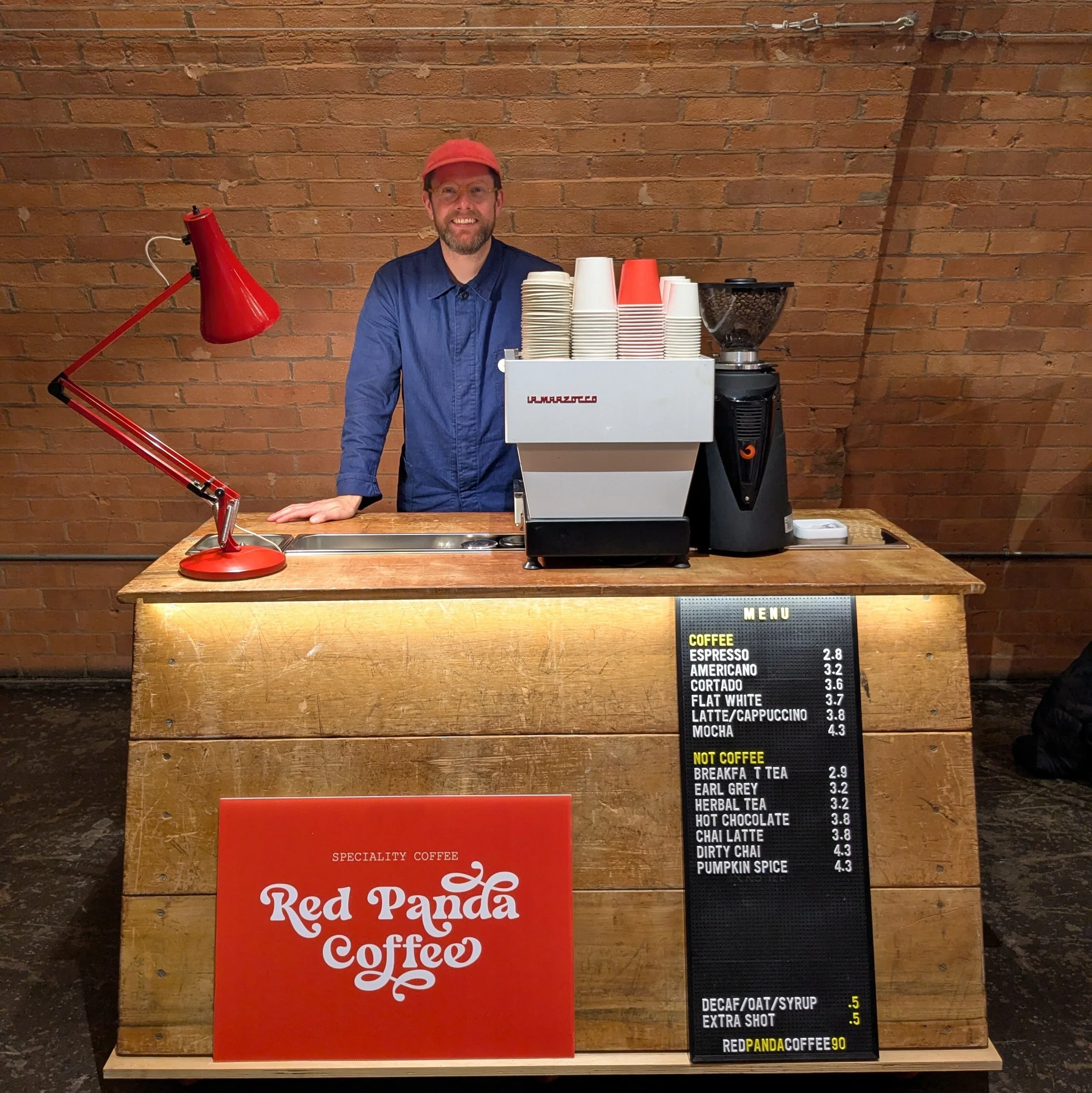A characterful Red Panda Coffee setup featuring a warmly lit wooden coffee bar, bold red branding and a sleek espresso machine, with a friendly barista standing behind the counter ready to serve. The industrial brick backdrop, retro red lamp and neat