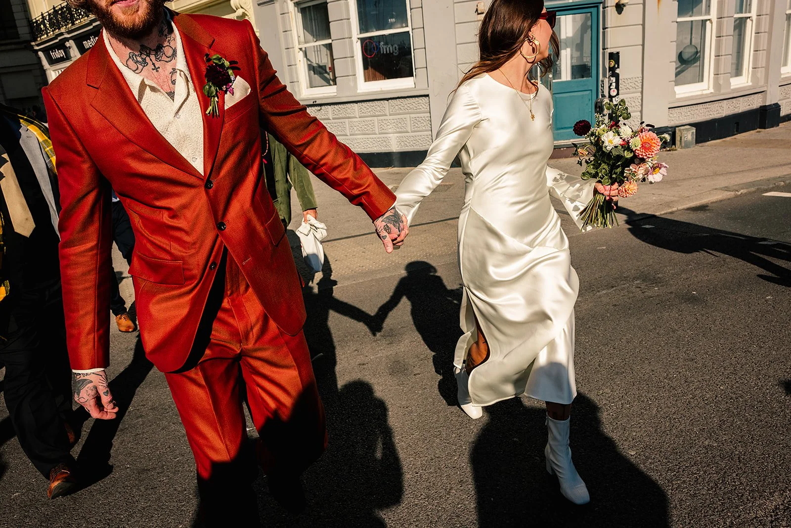 A stylish and modern wedding street-portrait captured by Chloe Mary Photo, showing a bride and groom striding confidently hand-in-hand through sunlit city streets. The groom wears a bold burnt-orange suit with a textured cream shirt and tattooed hand