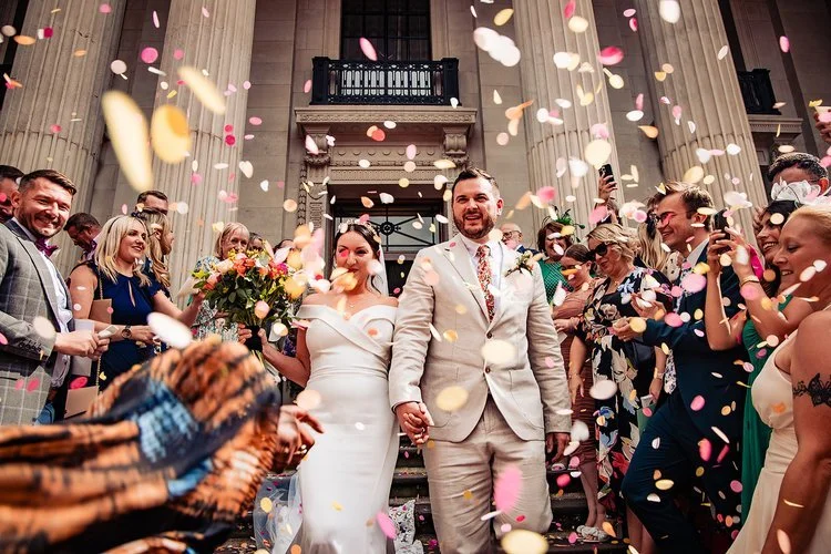 A vibrant and joy filled wedding moment captured by Natalia Case Photography, showing the couple walking hand in hand through a lively cascade of colourful confetti on the grand steps of a historic building. The groom’s light suit and patterned tie p