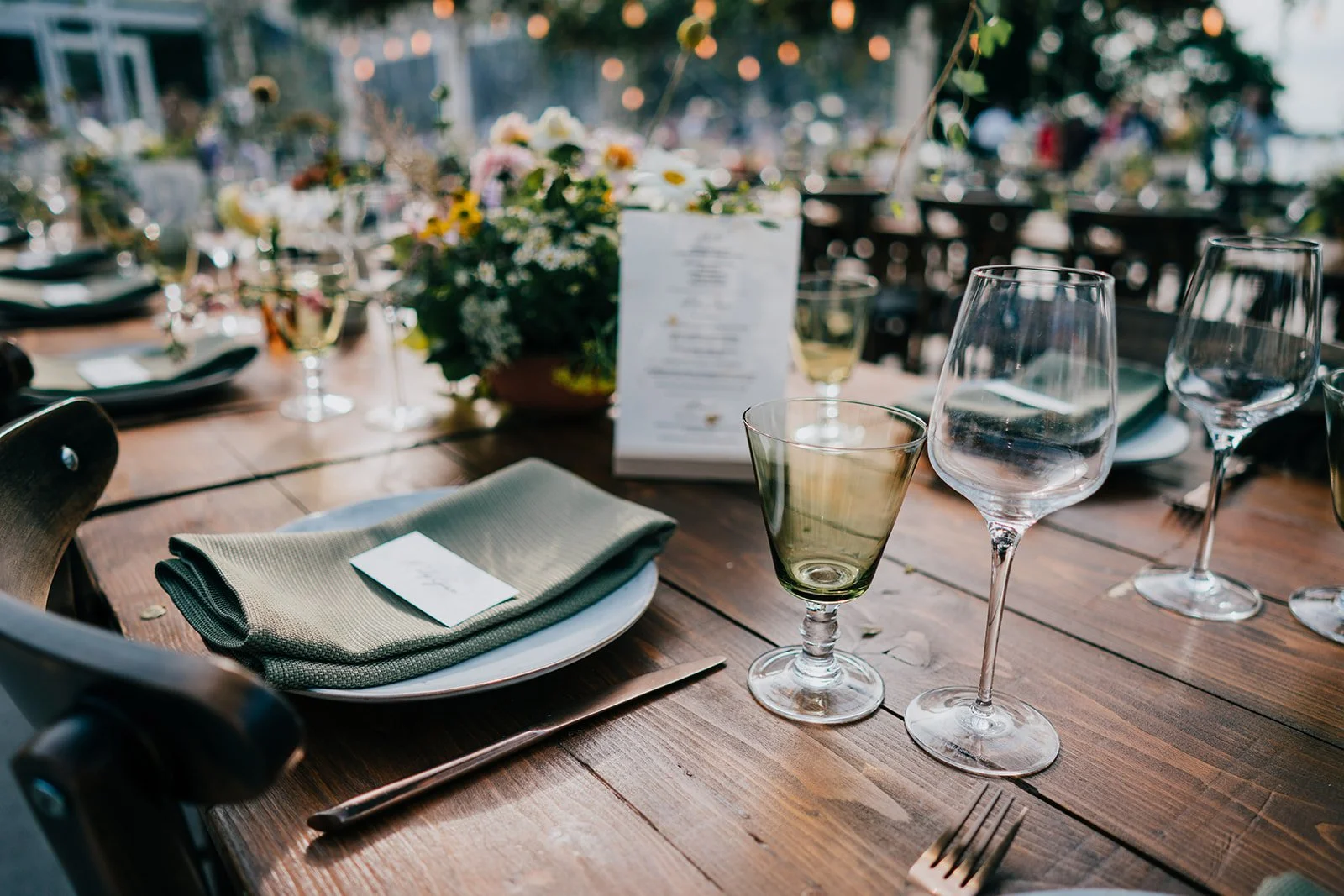 A beautifully styled rustic wedding tablescape curated by Keshira Creative Events, featuring wooden trestle tables set with sage-green linen napkins, elegant glassware, and modern cutlery. Fresh, wildflower-inspired centrepieces run down the length o