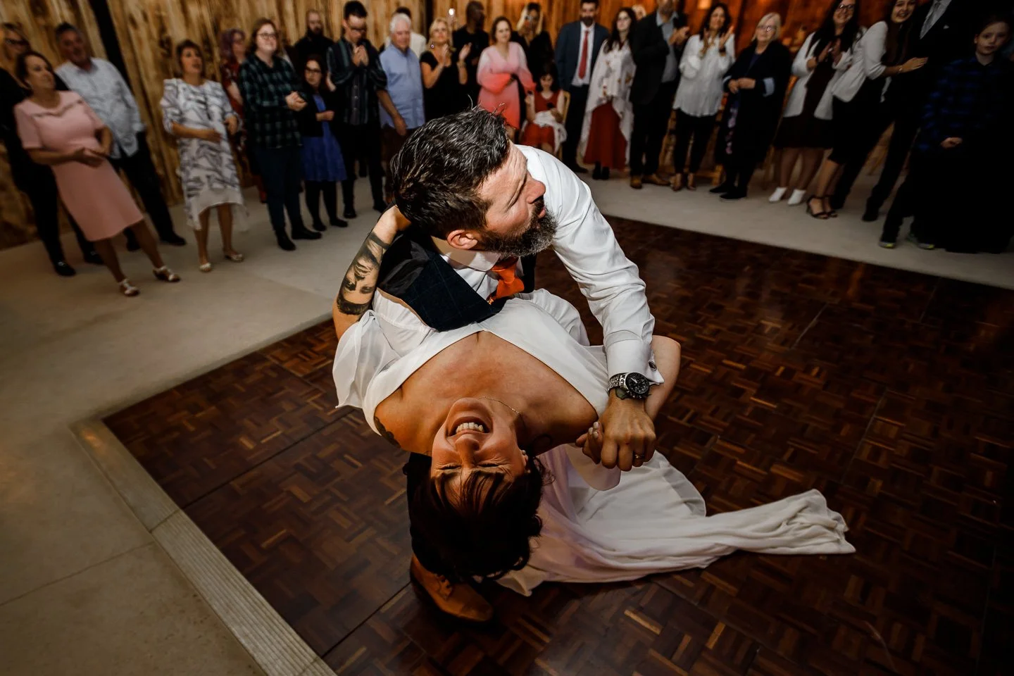 A joyful wedding first dance moment captured by Bluebell Photo Studio, showing the groom dramatically dipping his laughing bride on the dance floor while guests cheer around them. This lively candid wedding photo highlights natural storytelling, ener