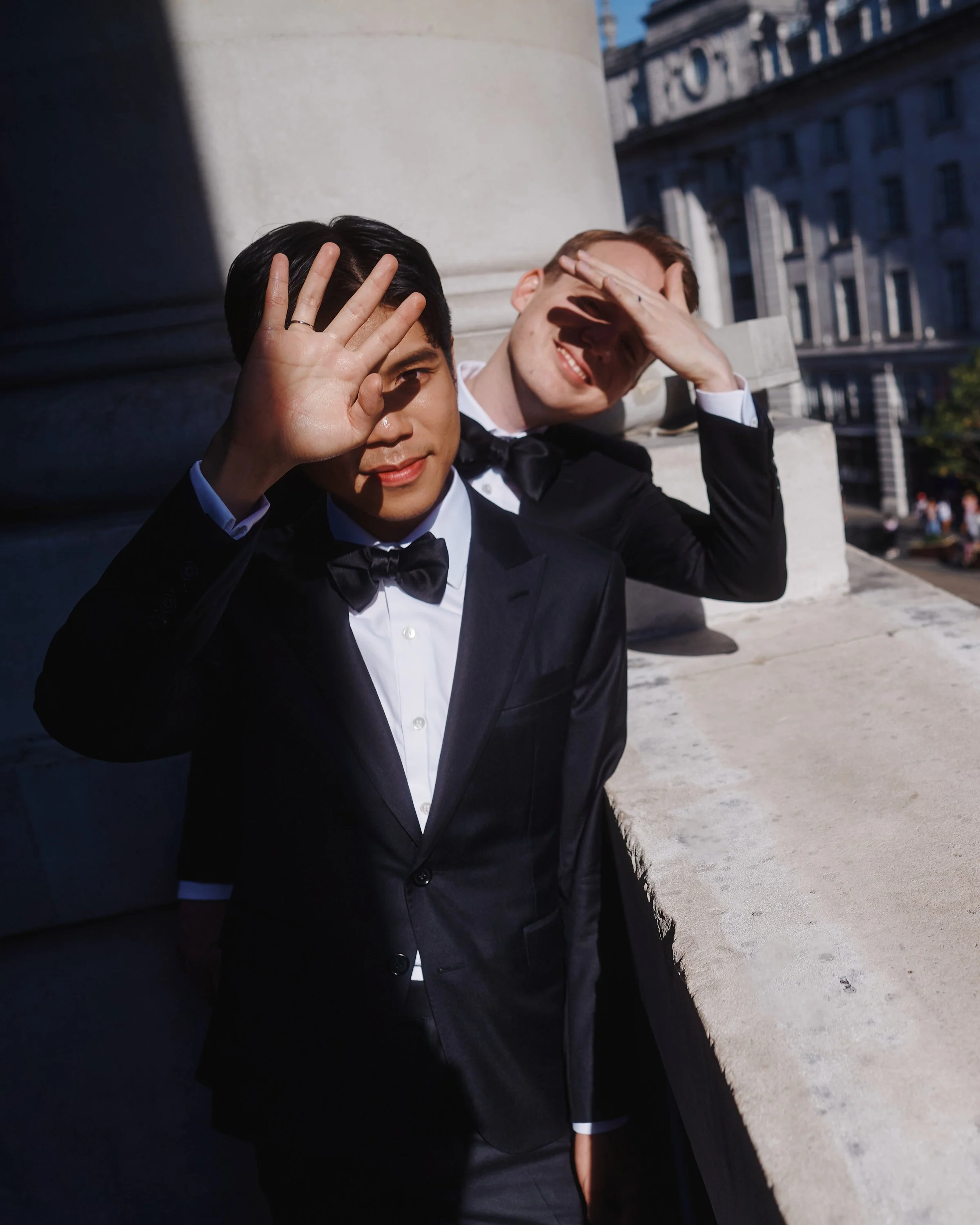 A bold and contemporary wedding portrait captured by Wayne La Photography, showing the couple playfully shielding their eyes from the bright afternoon sun. Their sleek black tuxedos and perfectly tied bow ties give the image a refined, modern edge, w