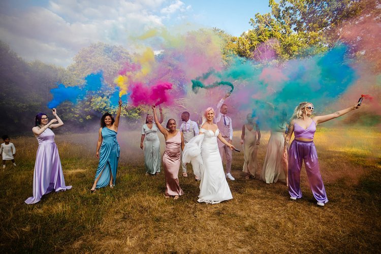 A vibrant outdoor wedding photo featuring a joyful bridal party walking across a sunny field while holding colourful smoke bombs in shades of blue, yellow, pink, and green. The bride in a white gown leads the group, surrounded by bridesmaids and groo