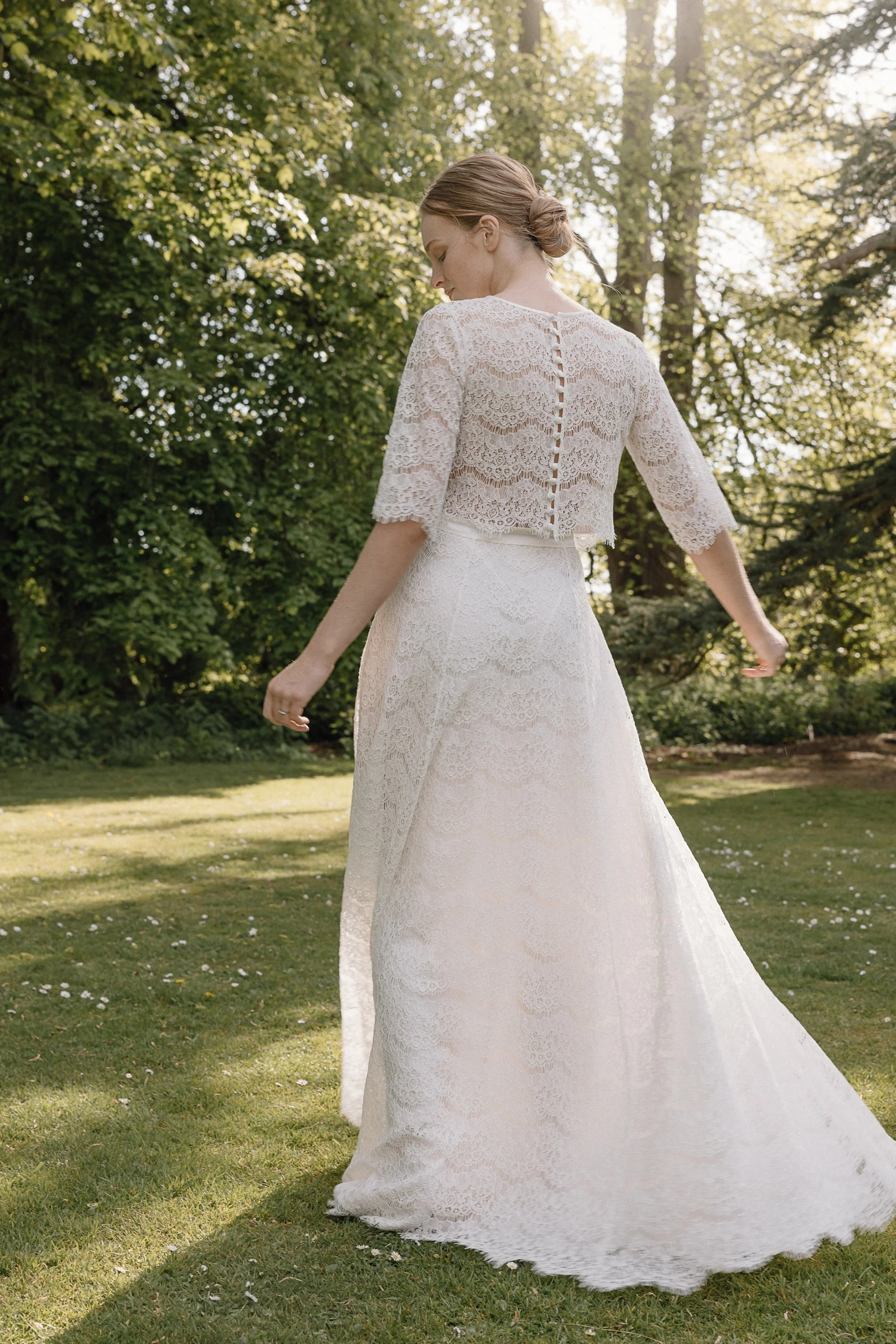 A bride walks across sunlit grass in an elegant lace two piece with scalloped detailing, the delicate buttons running down the back of her top catching the light while the surrounding trees cast soft dappled shadows that create a serene and romantic 