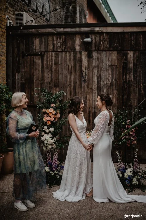 Two brides share a joyful moment during their outdoor wedding ceremony, beautifully officiated by modern celebrant Tab Taylor in a stylish sheer dress. Surrounded by romantic floral arrangements and rustic wooden backdrops, the intimate setting captu
