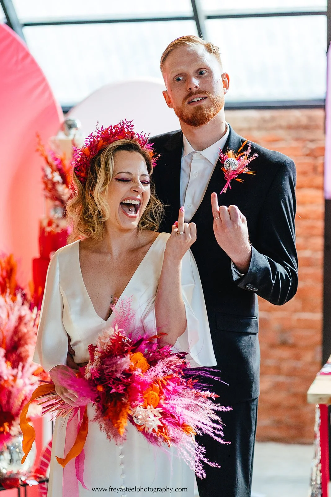 A joyful, rebellious portrait drenched in vibrant colour. The bride beams with laughter, wearing a bold pink dried-flower crown and holding a dramatic bouquet bursting with hot pink, orange, red and fuchsia stems, finished with feathery textures and 