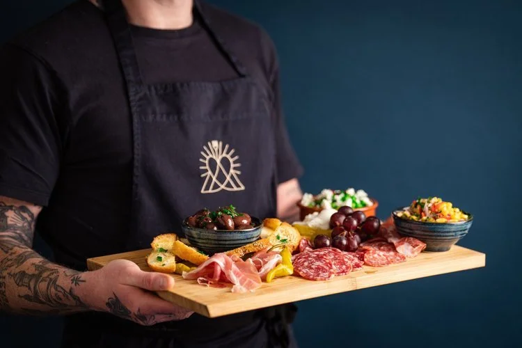 A Hardy’s Catering chef holding a rustic wooden grazing board filled with artisan cured meats, prosciutto, salami, marinated olives, crostini, grapes, and vibrant Mediterranean salads. Perfectly styled for luxury weddings and events, this charcuterie
