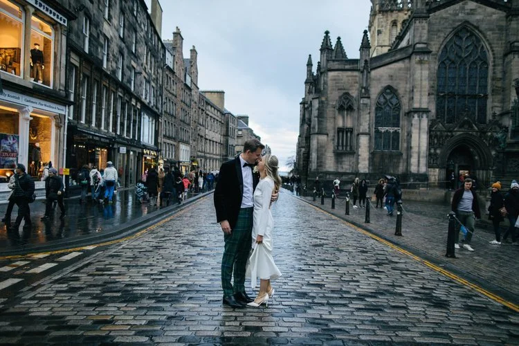 A romantic wedding portrait on Edinburgh’s iconic Royal Mile, photographed by Mirrorbox Photography. The couple stand together on the wet cobblestone street, surrounded by historic buildings and the dramatic façade of St Giles’ Cathedral. Passers-by 