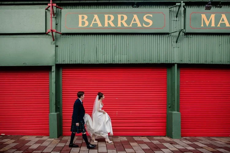 A vibrant, eye-catching wedding portrait captured by Mirrorbox Photography, featuring a couple walking past the iconic Barras Market in Glasgow. The bride lifts her dress slightly as she steps forward, her veil flowing behind her, while the groom in 