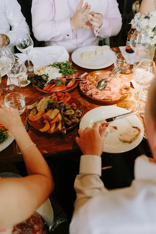 Guests enjoy a relaxed wedding meal featuring shared platters of roasted vegetables, cured meats, and fresh salads. The wooden table and family-style dining setup reflect Tasting Plates Wedding Caterers’ focus on sociable, seasonal, and beautifully p