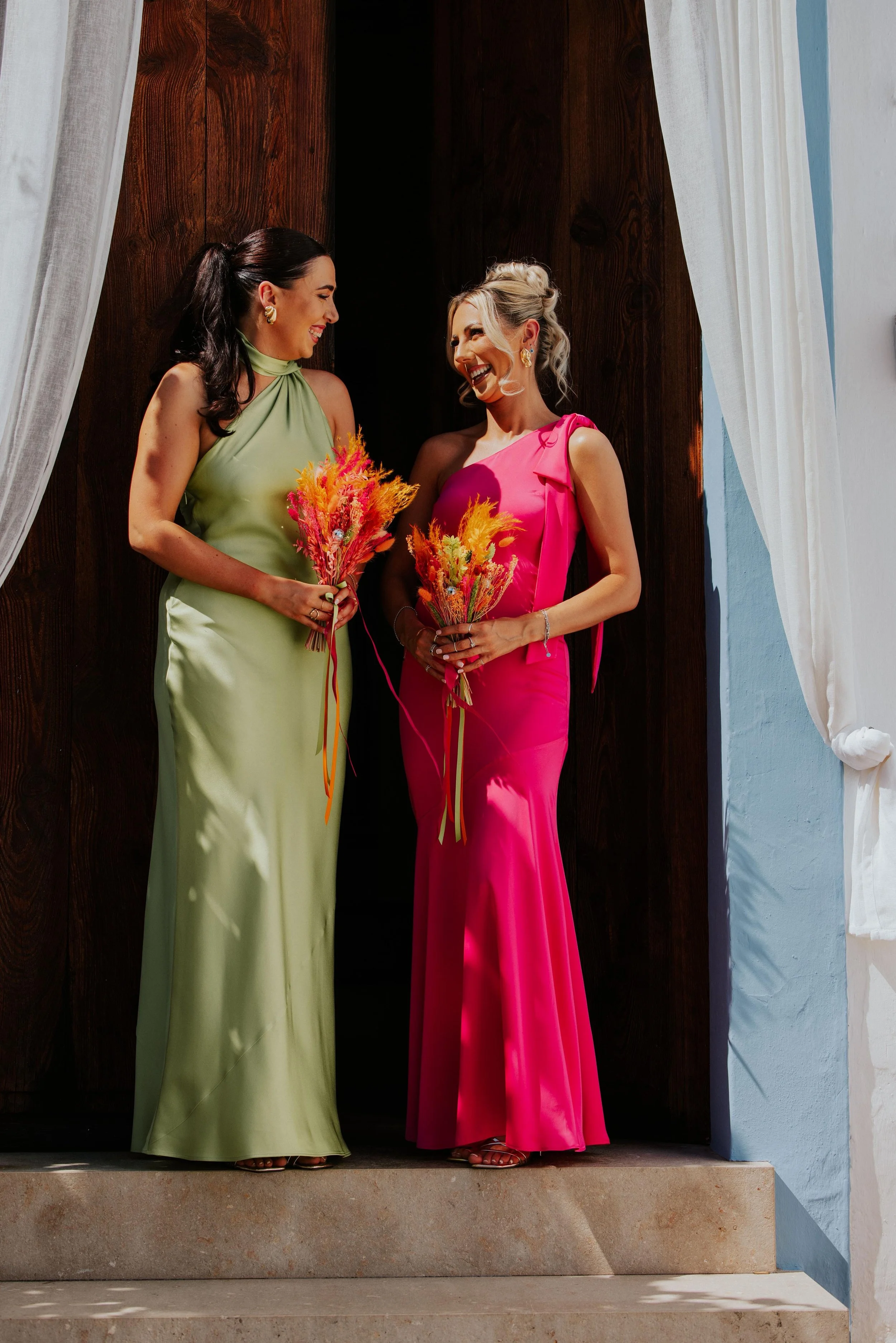 Two bridesmaids stand in bright sunlight against a dark wooden doorway, each holding a compact bundle of vivid dried stems in hot pink, orange and yellow, the bold colours of their bouquets echoing the striking contrast of their sleek satin dresses i