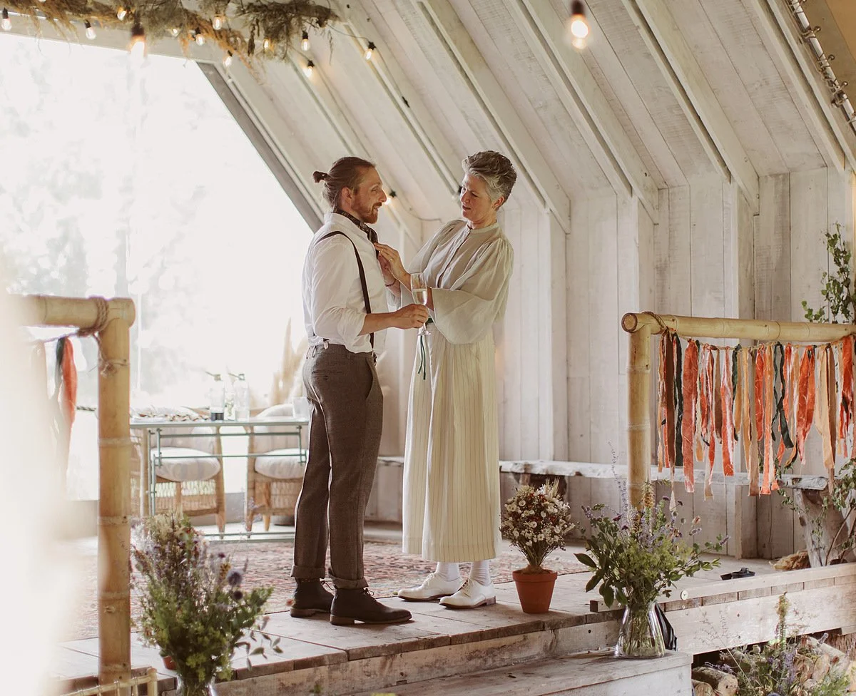 Arabella stands with one half of the couple on a raised wooden platform, gently adjusting their collar while holding a drink. The space is softly lit with natural light and simple floral details. Nick Ray Photography.
