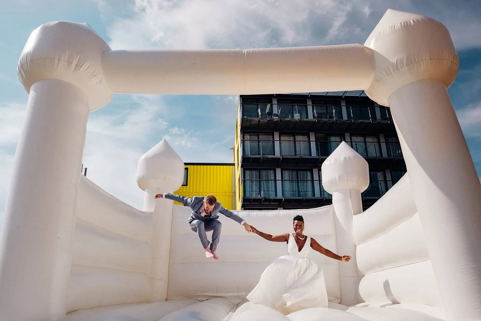 Two people in wedding attire are holding hands and bouncing on a large white inflatable castle outdoors, with one wearing a grey suit mid-jump and the other in a white dress smiling as they land. The bright scene is set against a blue sky with light 