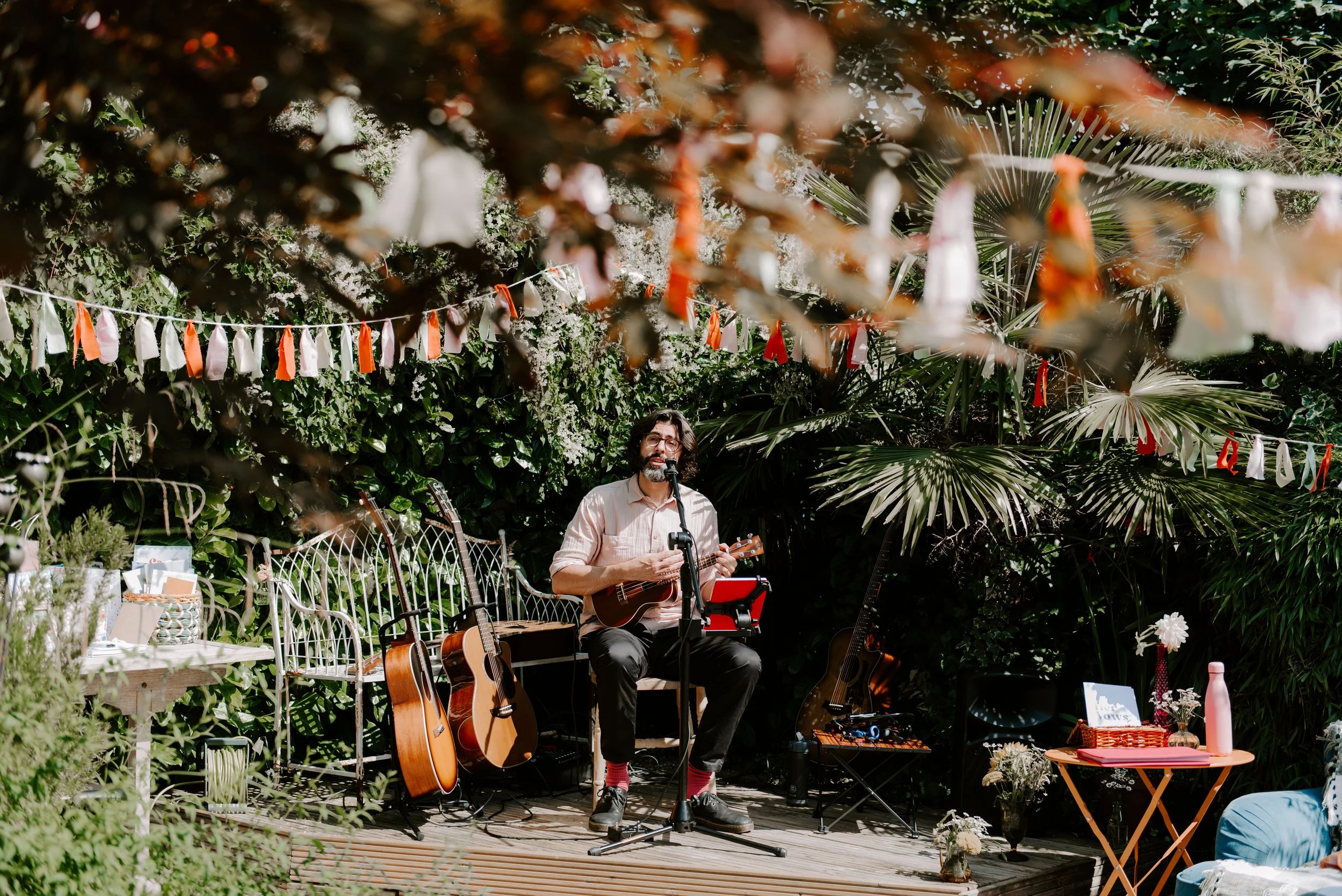 Thomas J Wilman, an acoustic wedding singer and guitarist, performs live in a garden setting surrounded by vibrant greenery and acoustic guitars. His relaxed and natural style creates a warm, intimate atmosphere perfect for outdoor weddings and laid-