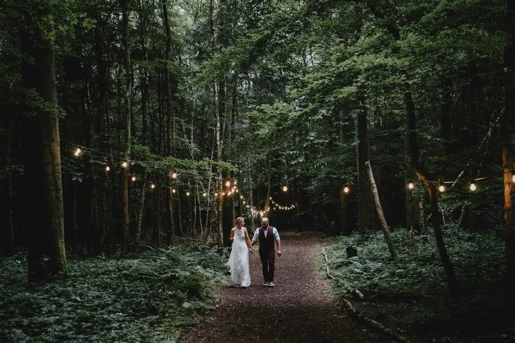 A magical and atmospheric woodland wedding portrait captured by Matt Fox Photography, showing a couple walking hand in hand along a forest path lined with glowing string lights. The tall trees and dense greenery create an immersive, fairytale like se