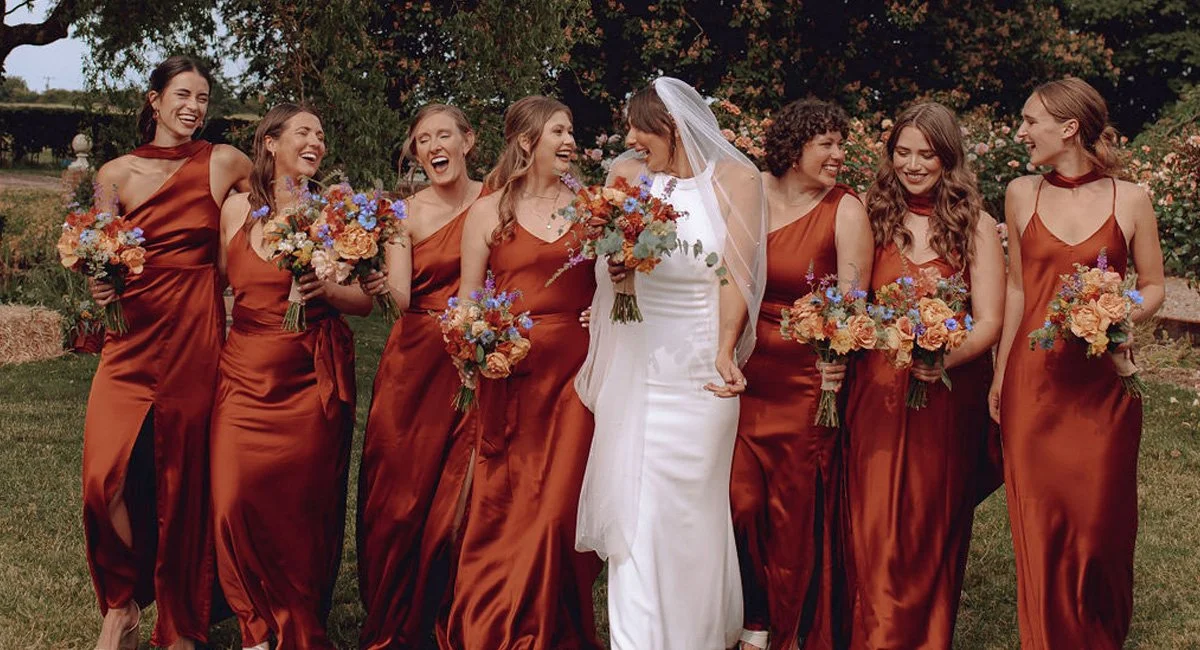This joyful bridal party showcases Rewritten Bridal Wear’s burnt orange satin bridesmaid dresses, a warm and modern take on autumn wedding style. Each gown features a slightly different silhouette - from asymmetric necklines to strappy cowl designs -