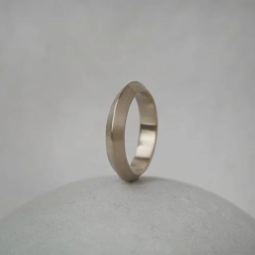 A minimalist 18ct white gold knife-edge wedding band by Natalie Vernazza, featuring a sleek apex profile and polished finish, photographed standing upright on a soft neutral background to highlight its modern, sculptural design.