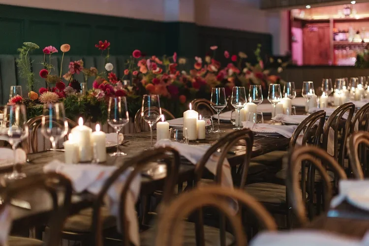 Romantic candlelit wedding table setup at Colours Hoxton in Shoreditch, London. The intimate dining scene features rustic wooden tables lined with glowing white candles, elegant glassware, and lush floral arrangements in rich pink, red, and coral ton
