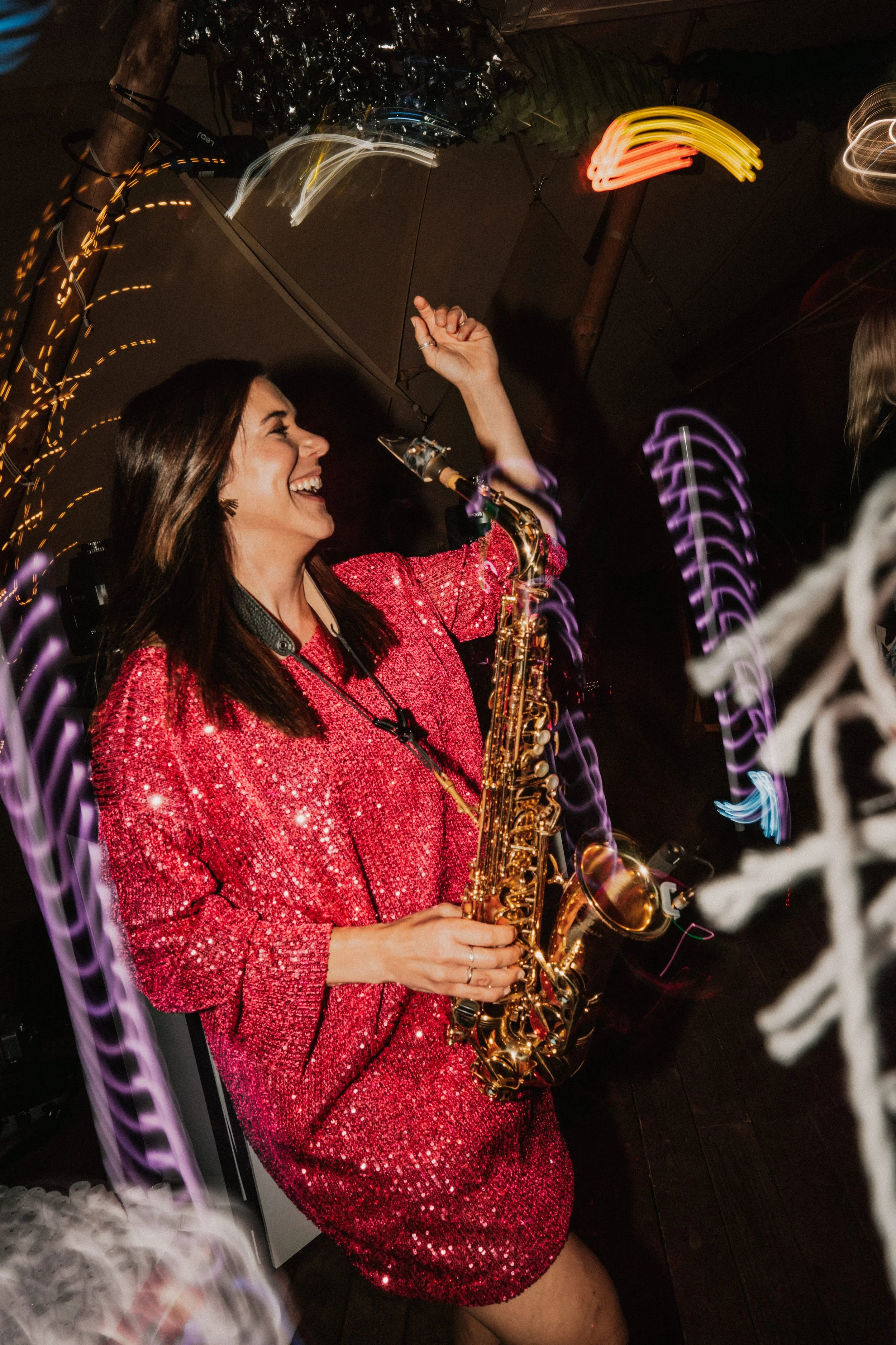 A vibrant wedding saxophonist from Married to the Music performs live during a lively evening reception, wearing a sparkling pink sequin dress and surrounded by colorful party lighting. The image captures the joy and energy of live saxophone music fo