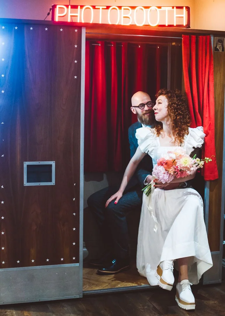 A playful and stylish wedding portrait captured by Barbara K Photography, featuring a couple sitting together in a retro photobooth under a glowing neon sign. The bride, wearing a ruffled white dress paired with chunky platform shoes, holds a colourf