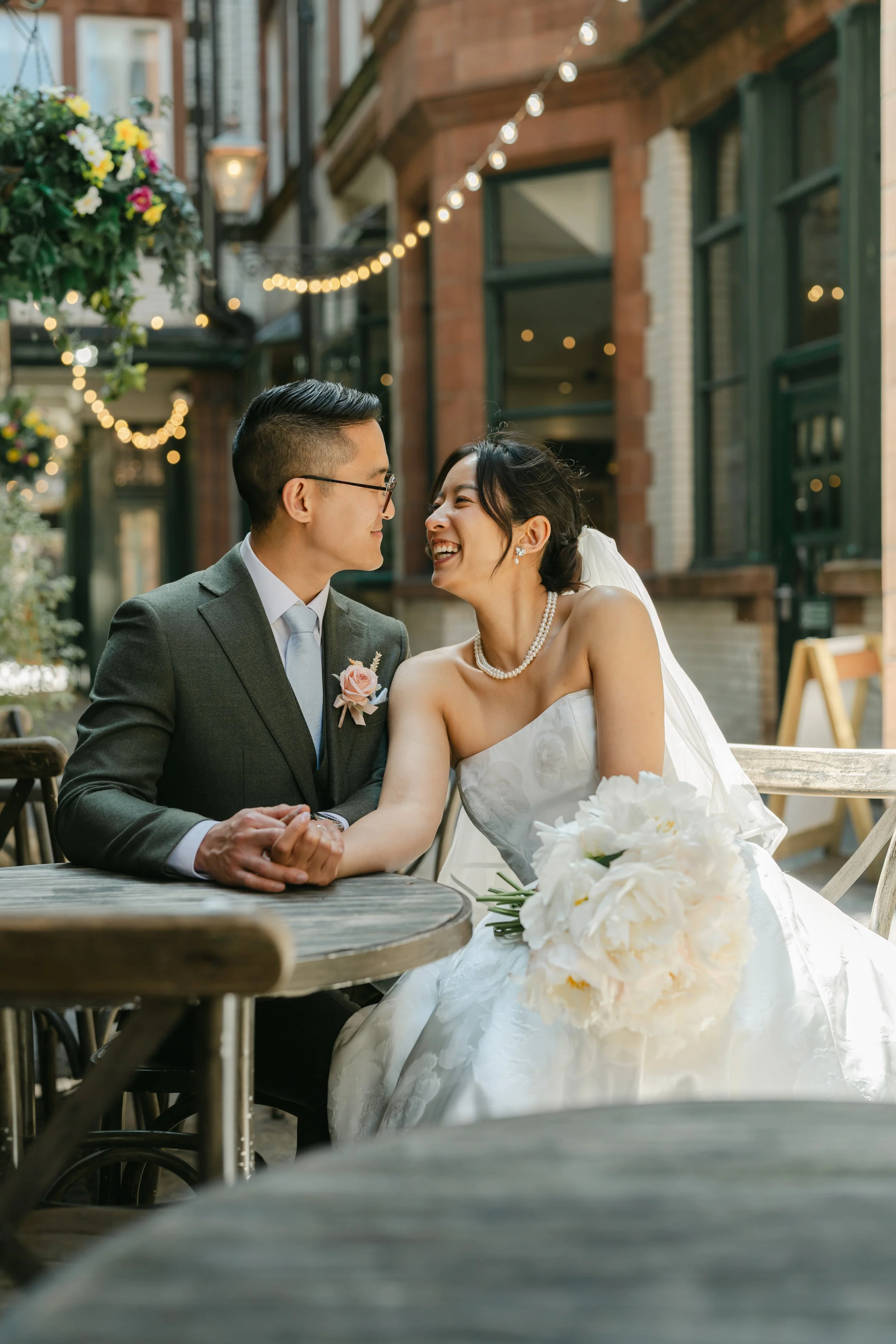 A bride and groom sit together at an outdoor café table, sharing a joyful moment in a charming city street beautifully captured by Christy Photography. Warm string lights, hanging flowers and historic brick buildings frame the couple as they hold han
