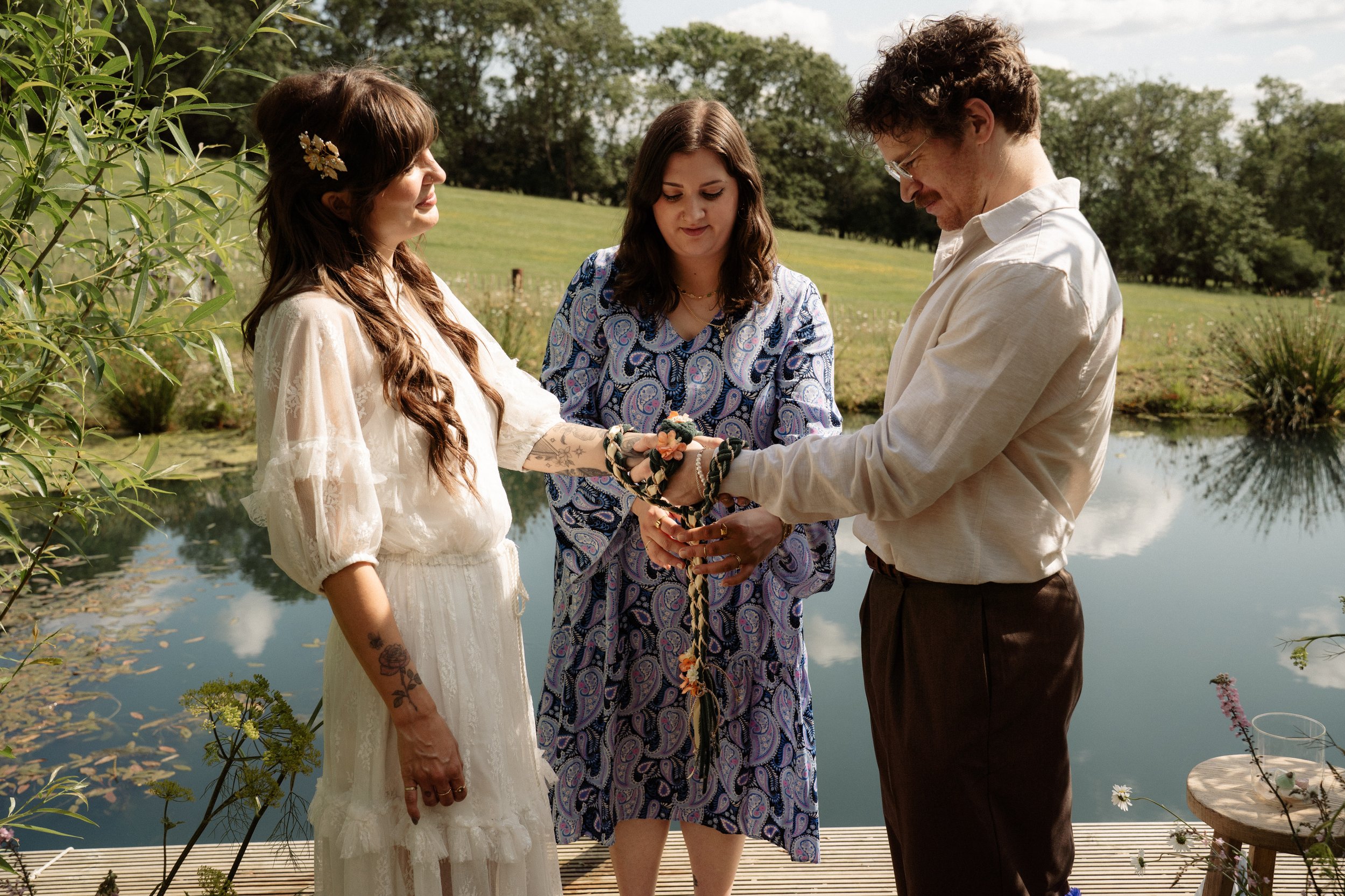 A touching moment during an outdoor wedding ceremony. The couple stands facing each other beside a peaceful pond, their hands bound together in a hand-fasting ritual - a traditional symbol of unity and love. Lauren The Spiritual Celebrant, standing b