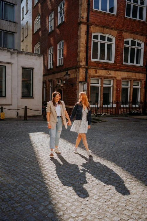 A couple walk hand in hand through a sunlit cobbled street, captured by Joshua Humphrey Photography. The warm light and their relaxed smiles create a natural romantic portrait set against charming historic city architecture.