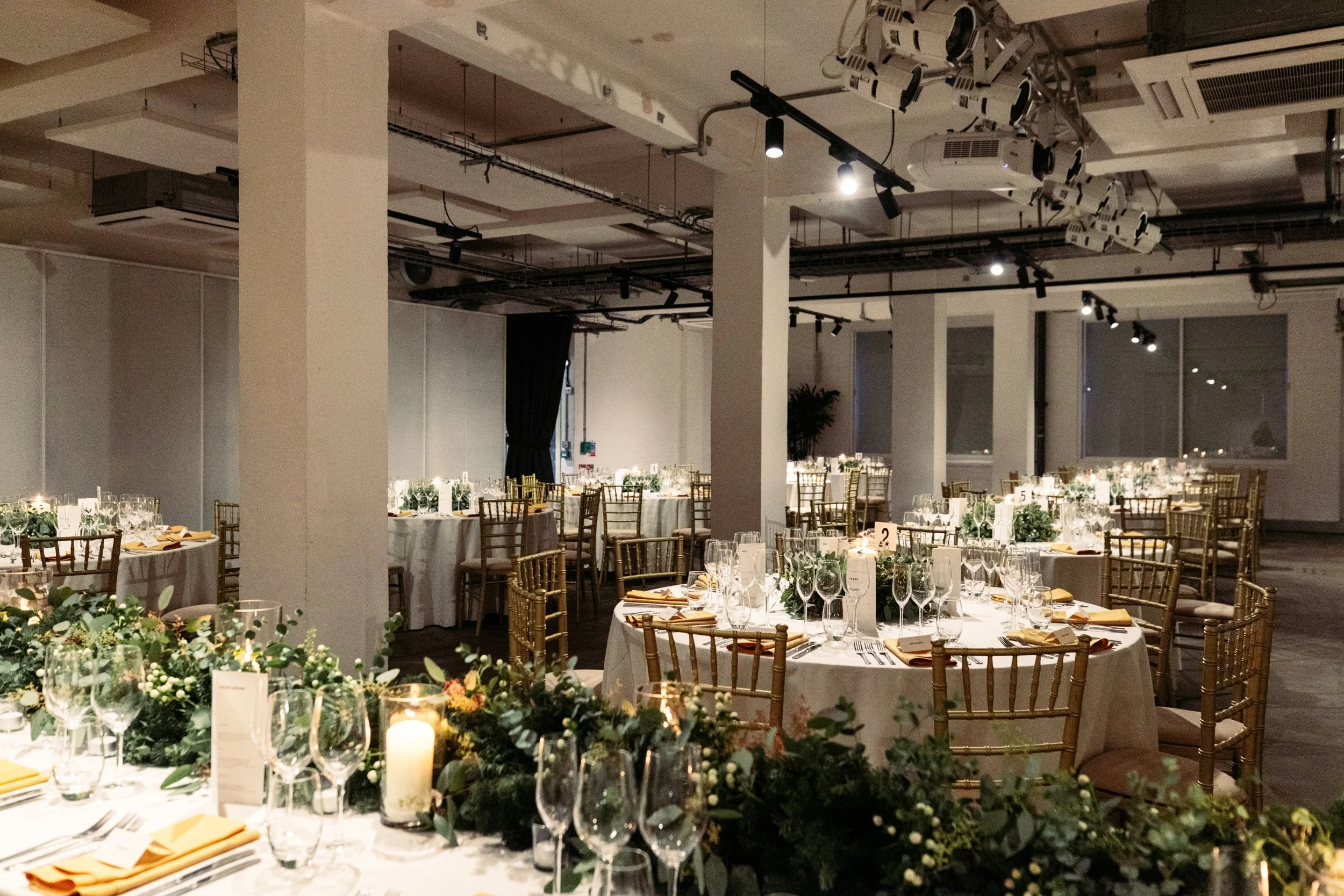 An elegant wedding reception setup at Ministry Venues, featuring round tables with crisp white linens, gold Chiavari chairs, candlelit greenery centrepieces and soft ambient lighting, creating a refined yet contemporary atmosphere for modern London w