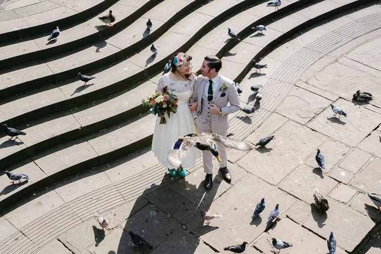 A couple walk arm in arm across a sunlit stone amphitheatre surrounded by pigeons, captured beautifully by Christy Photography. The elevated viewpoint, curved steps and movement of the birds create a dynamic, city-inspired backdrop that highlights th