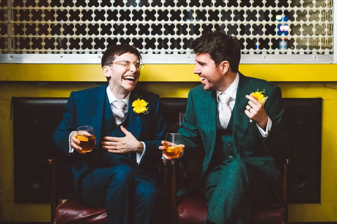 A relaxed and joy-filled moment between two grooms captured by Barbara K Photography, as they sit together laughing over celebratory drinks. Both wear stylish three-piece suits with bright yellow boutonnieres that add a pop of colour against the mode