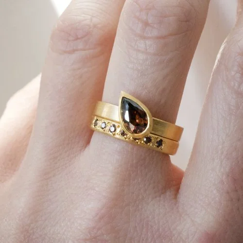 A pear-shaped cognac diamond engagement ring paired with a matching yellow gold eternity band, featuring warm-toned diamonds and a modern, handcrafted design by Natalie Vernazza, worn on the hand to show scale and detail.
