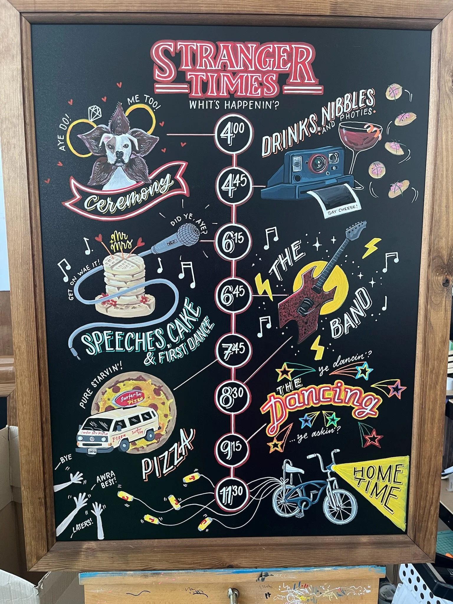 A hand-illustrated wedding timeline sign is displayed in a wooden frame, designed in a bold chalkboard style with vibrant colours and playful lettering. The layout runs vertically, marking key moments of the day with illustrated icons including a cer