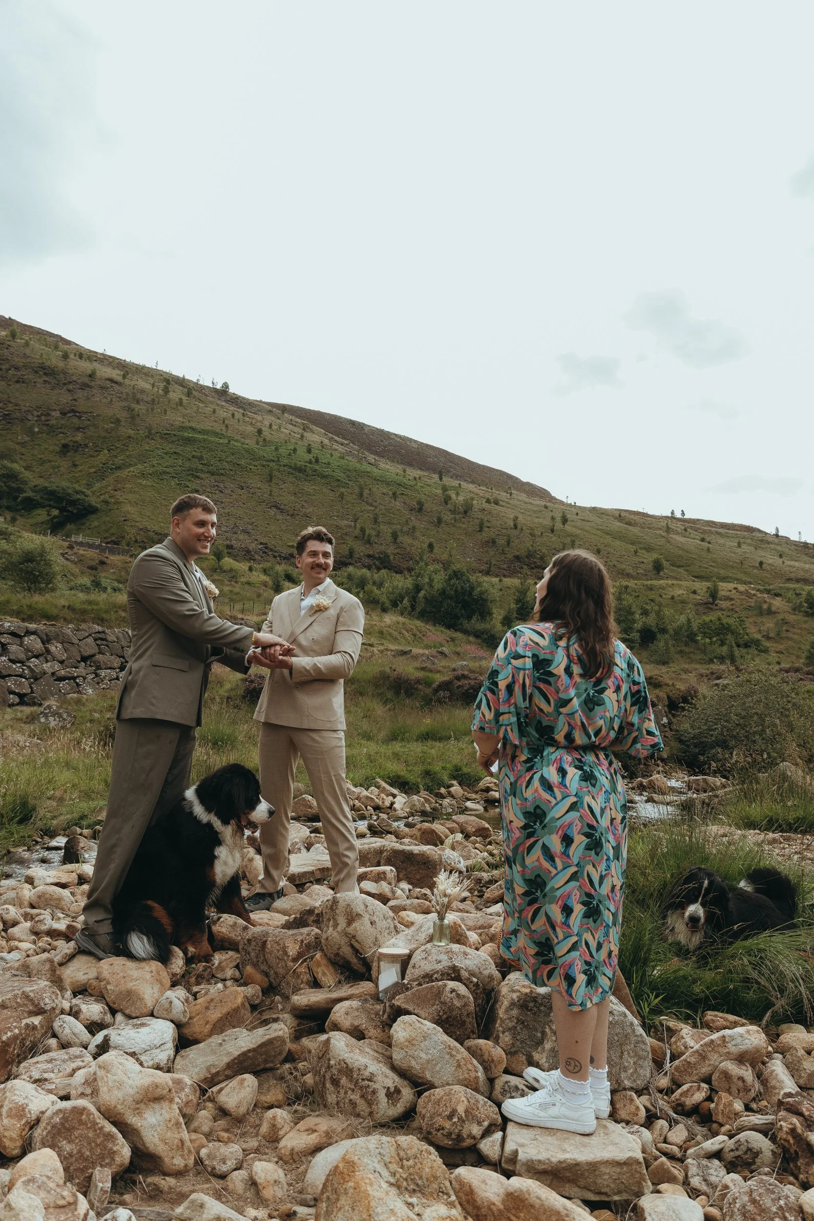A beautiful outdoor wedding ceremony set in a rugged, natural landscape. Two grooms stand hand in hand on a rocky riverbank, smiling warmly at each other. Lauren The Spiritual Celebrant faces them, holding a book, dressed in a colourful patterned dre