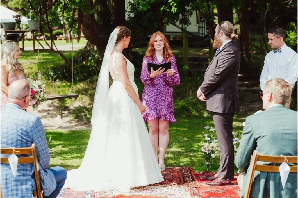 Outdoor summer wedding ceremony conducted by celebrant Meg Senior in a pink and purple dress, standing between the bride and groom under the trees. The couple exchange vows surrounded by friends and family seated on wooden chairs, with boho-style rug