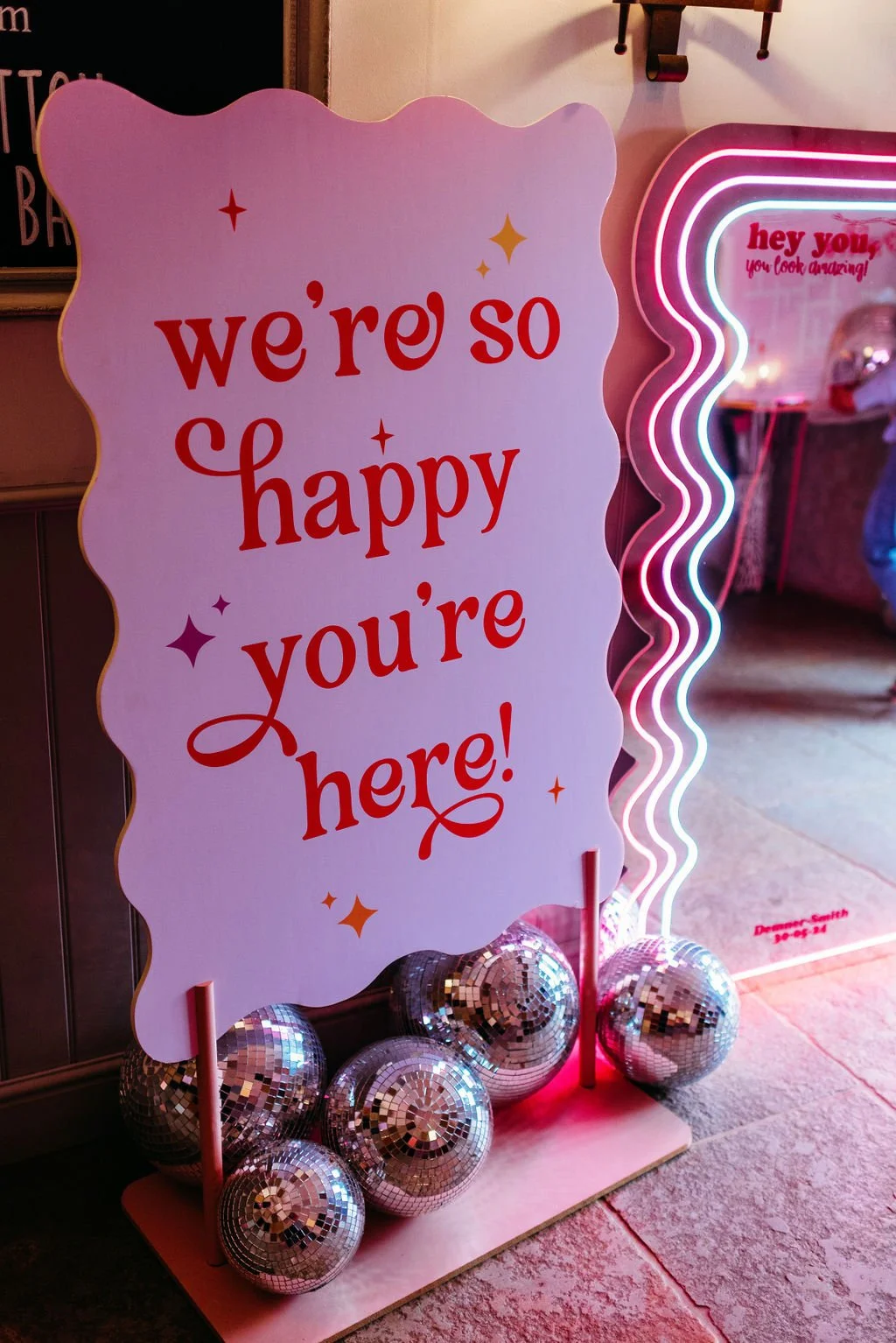 Playful wedding welcome sign by AMW Events reading “we’re so happy you’re here!” in red retro-style font, surrounded by sparkling disco balls, next to a neon-lit wavy mirror in a vibrant pink setting.