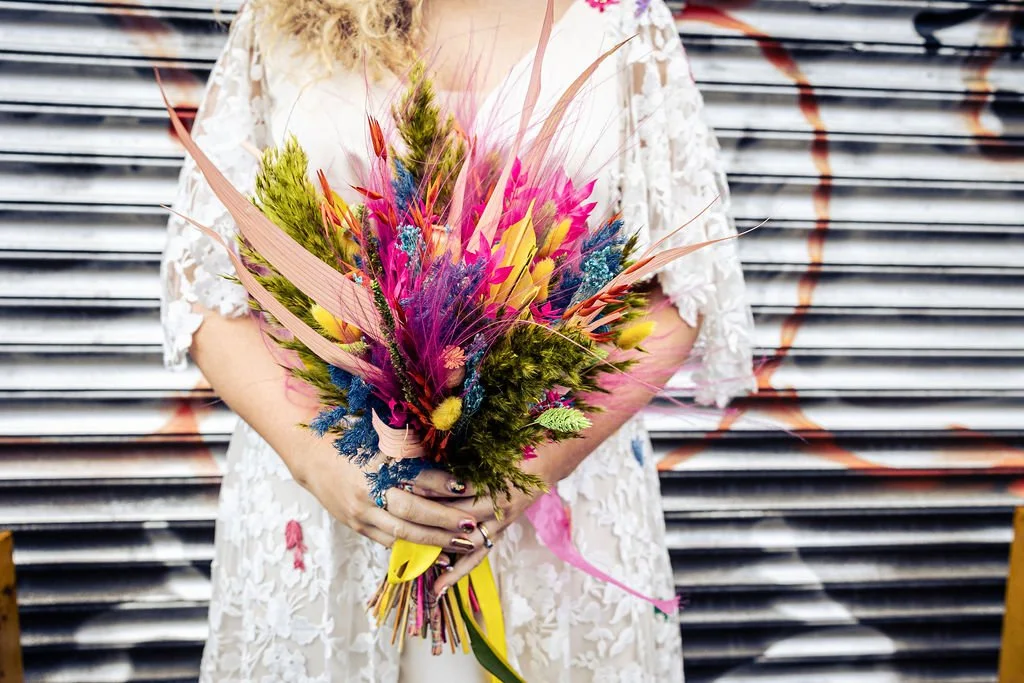 The bride holds a striking dried flower bouquet bursting with vivid colour, the neon pinks, electric blues, sunny yellows and fresh greens creating a bold contrast against her lace dress while feathery textures, spiked stems and painted foliage lend 