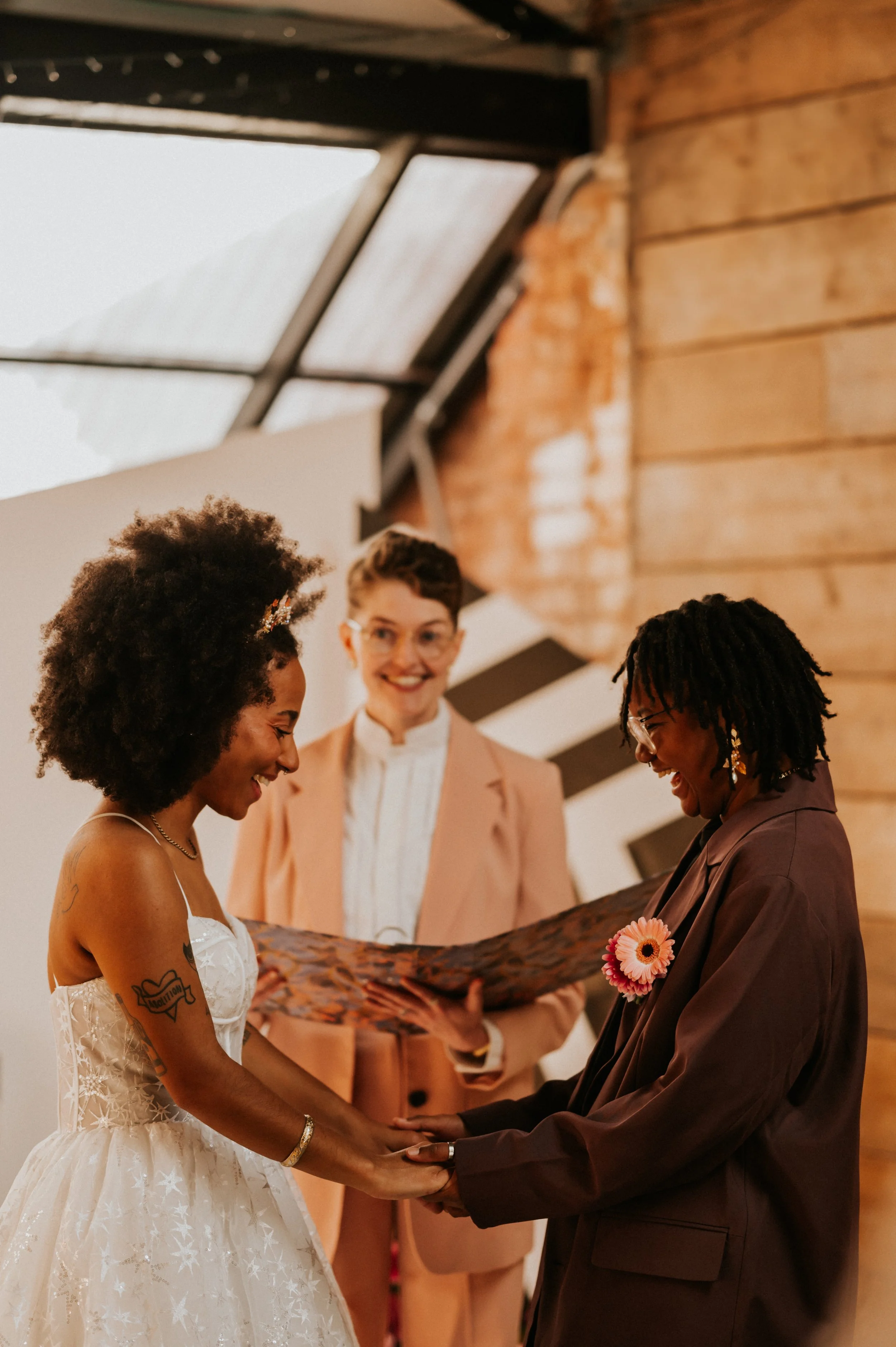 A wedding ceremony in the UK, featuring celebrant Chloe Green officiating as two brides exchange vows with love and laughter. One bride wears a sparkling white gown, the other a chic tailored suit, creating a beautiful blend of individuality and conn