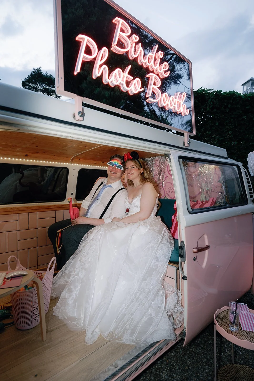 The couple sit tucked inside a pastel pink photo booth van surrounded by shimmering tinsel and playful props while a glowing neon sign above them lights up the evening sky and adds a fun celebratory feel to the moment. Tash Busta Photography. 