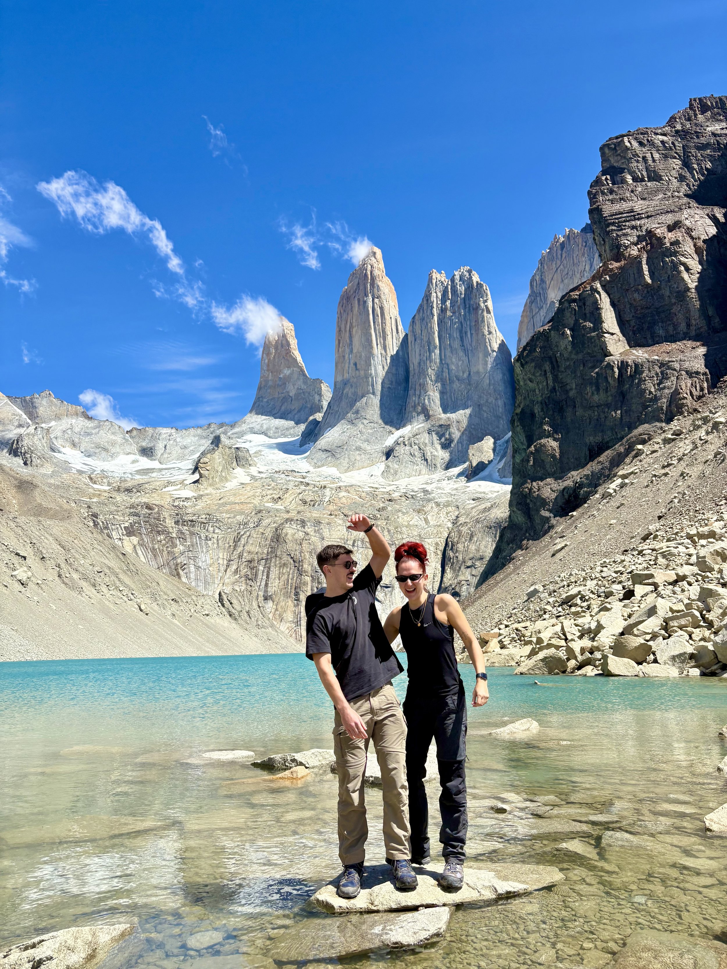 Robyn Smith Wedding Celebrant stands with a friend on stepping stones in a turquoise glacial lake, framed by dramatic mountain peaks and bright blue skies.