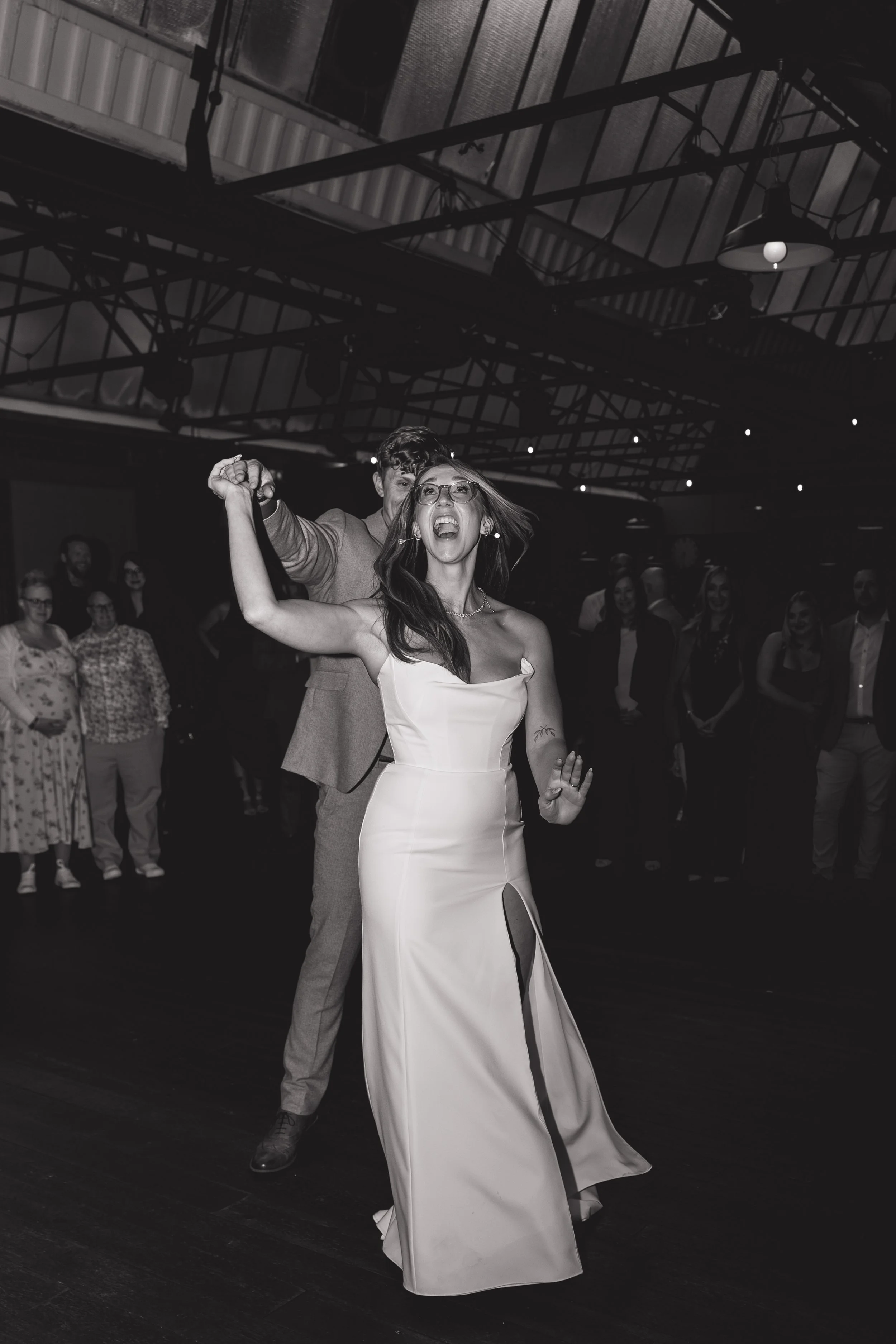 A vibrant and emotional first dance moment captured by Joy Louise Photography, showing the bride and groom laughing as they move across the dance floor surrounded by their guests. The bride leads with infectious joy, her fitted satin gown catching th