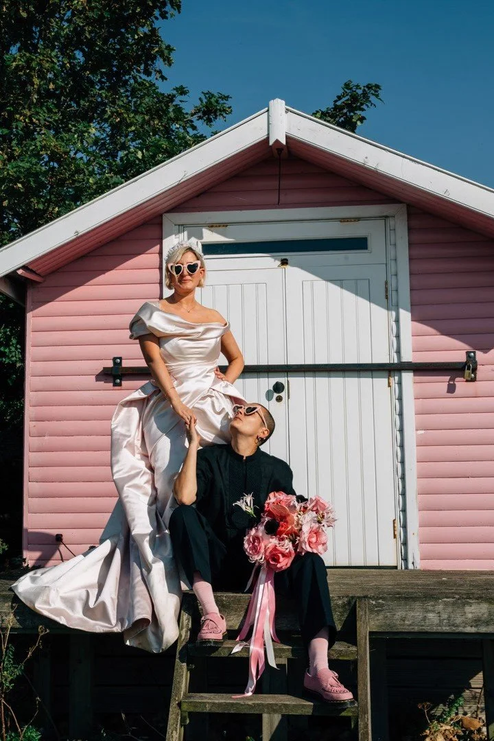 A stylish wedding couple strike a bold, fashion-forward pose outside a pastel-pink beach hut in bright summer sun. One partner stands confidently on the steps in black tailoring, pink socks and loafers, holding a vibrant pink bouquet with long ribbon