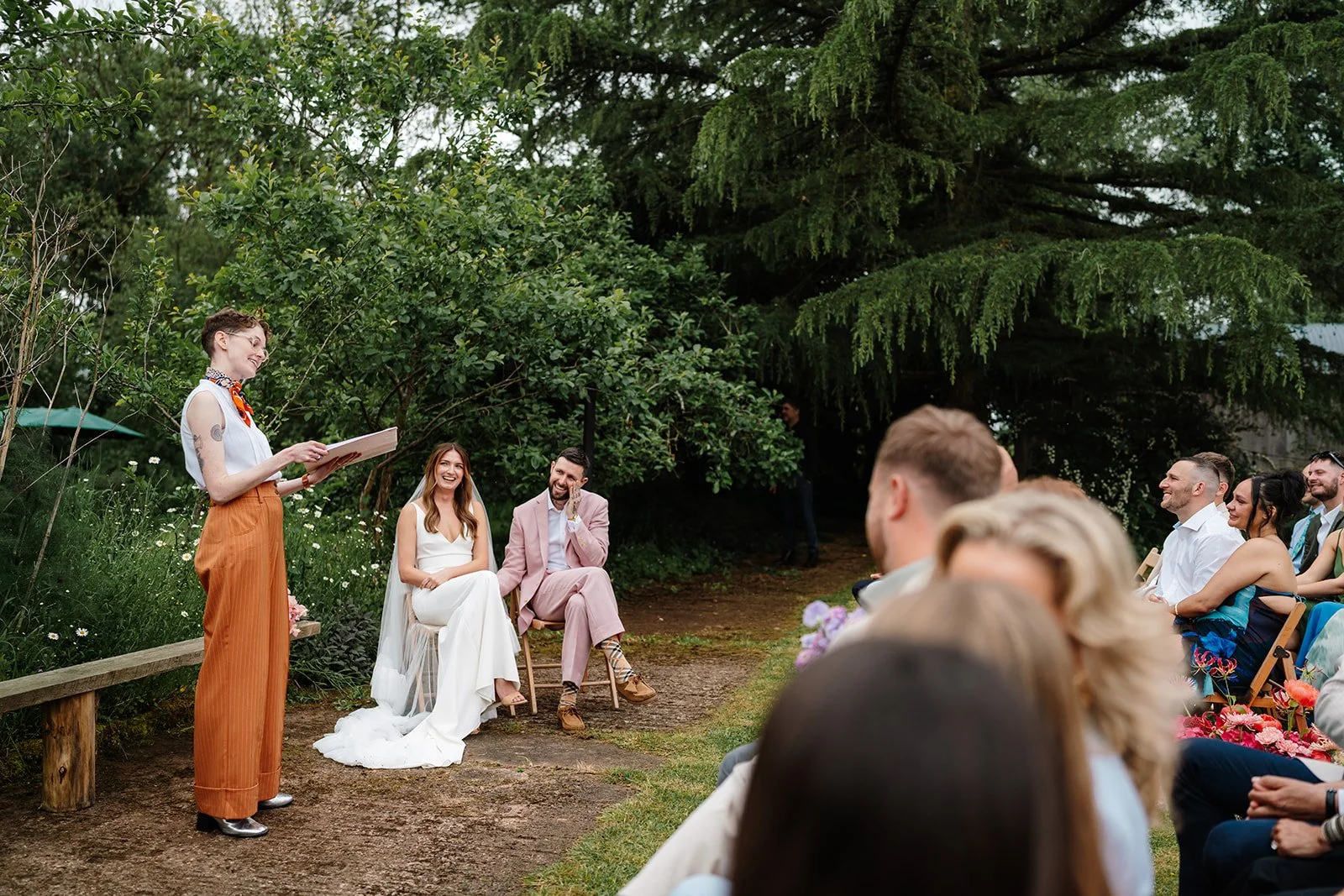 An outdoor wedding ceremony led by UK celebrant Chloe Green dressed in stylish rust trousers and a white shirt, delivering a heartfelt ceremony to a laughing couple and their guests. Set against a lush green backdrop, the bride and groom sit together