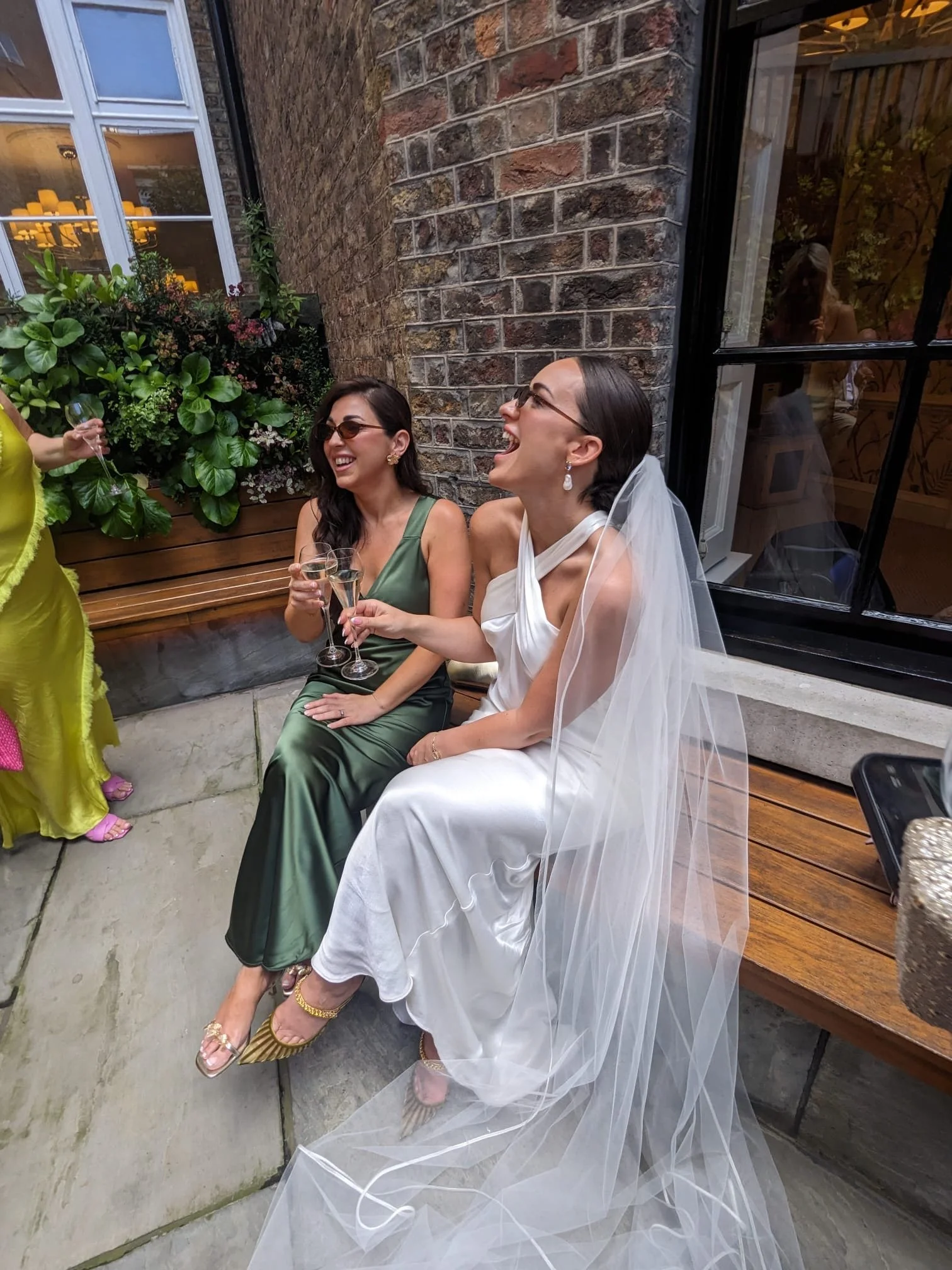 A joyful candid featuring Rewritten Bridal Wear - the perfect example of their modern, minimalist bridal and bridesmaid style. The bride wears a sleek satin gown with a draped neckline and halter-style wrap detail, paired beautifully with a long, flo