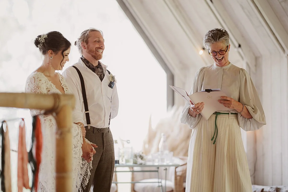 Arabella stands facing the couple, reading from her notes with a broad smile, while they stand side by side on the platform, hands joined and looking towards her. Soft daylight fills the space, with simple wooden details and light fabrics framing the