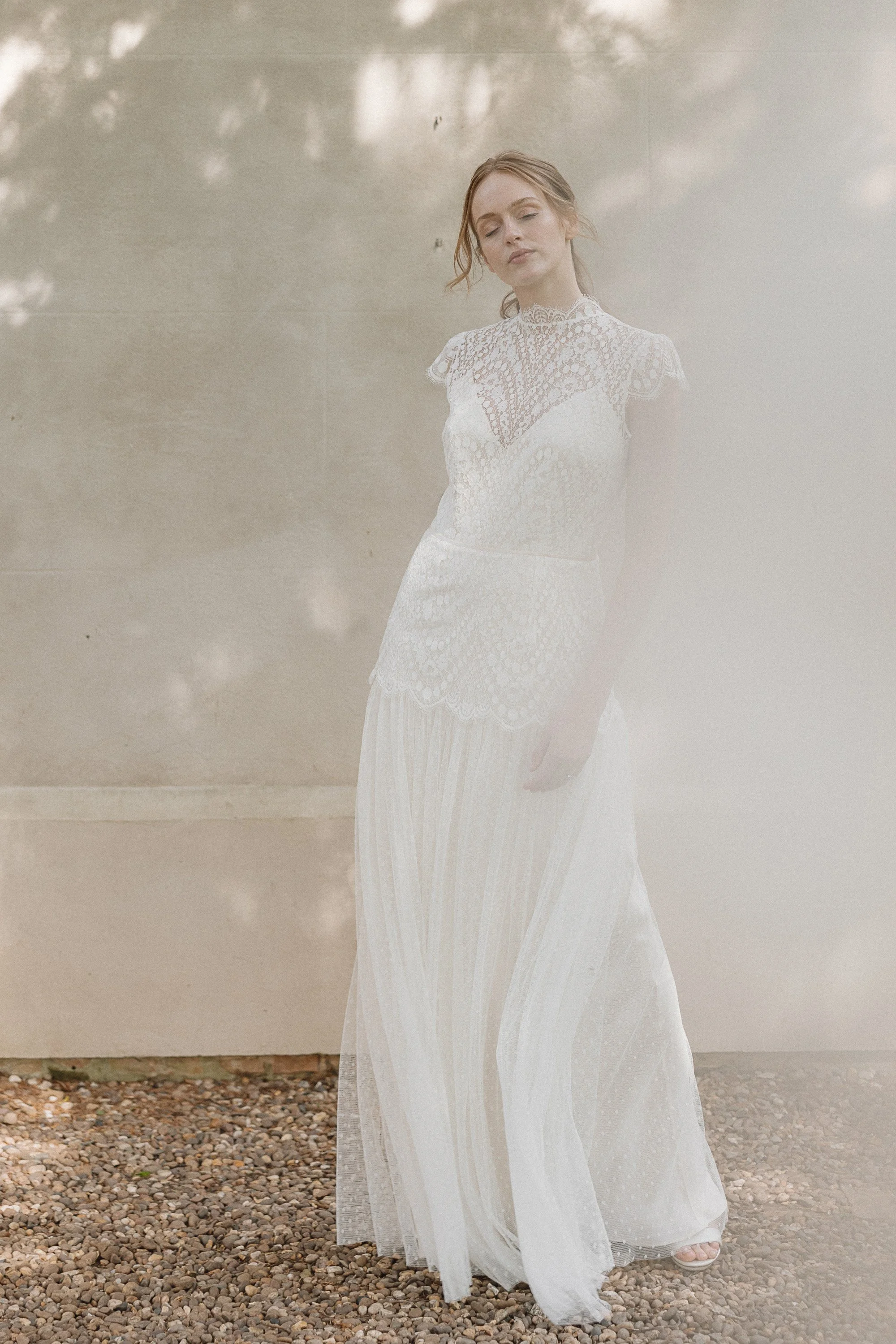 Soft light filters through the trees as the bride stands against a pale stone wall wearing a high neck lace gown with delicate cap sleeves and a scalloped bodice that flows into a sheer dotted skirt which moves gently around her feet creating an ethe