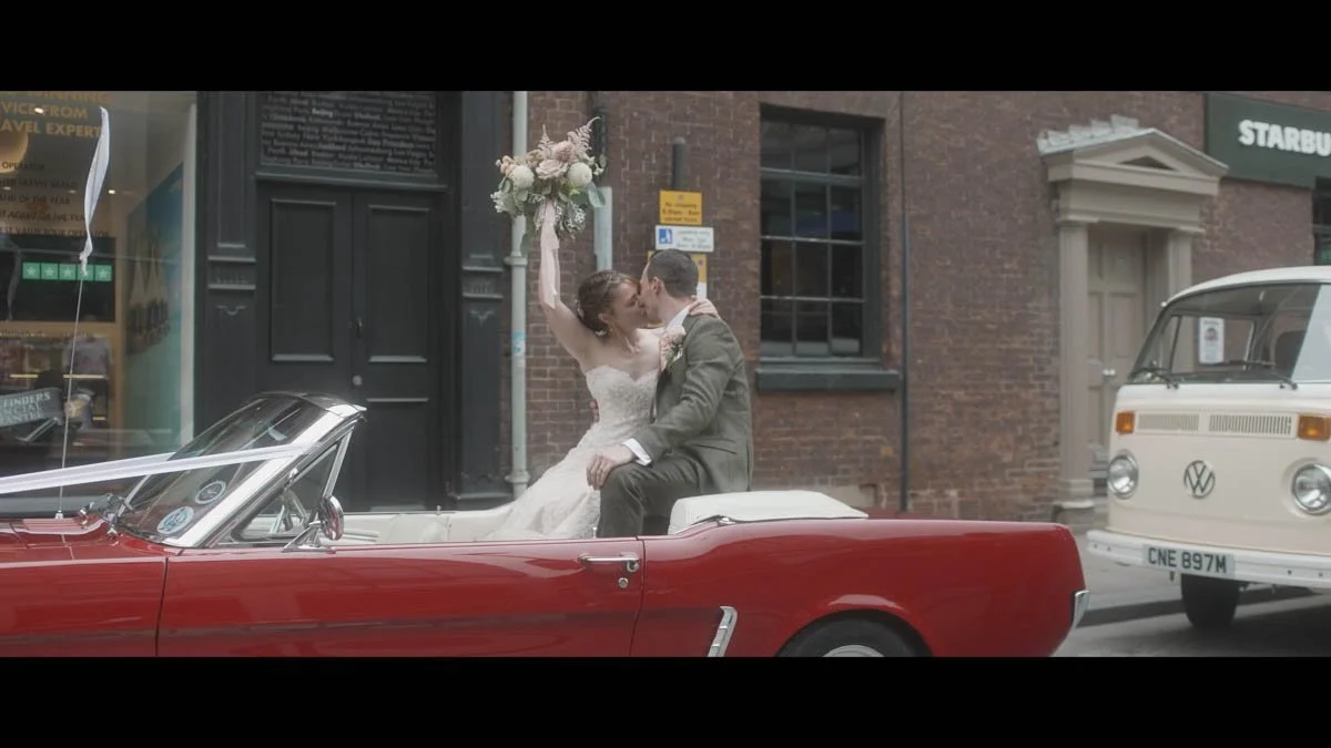 A newly married couple shares a joyful kiss while perched in a glossy red convertible with the bride lifting her bouquet triumphantly into the air as soft light settles over the brick lined street and a vintage cream Volkswagen nearby adding to the c