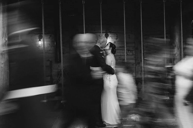 A striking and atmospheric wedding portrait captured by YU Photography, showing the couple sharing an intimate kiss while guests sweep past them in a blur of motion. The black and white tones enhance the drama, drawing the eye to the stillness and co