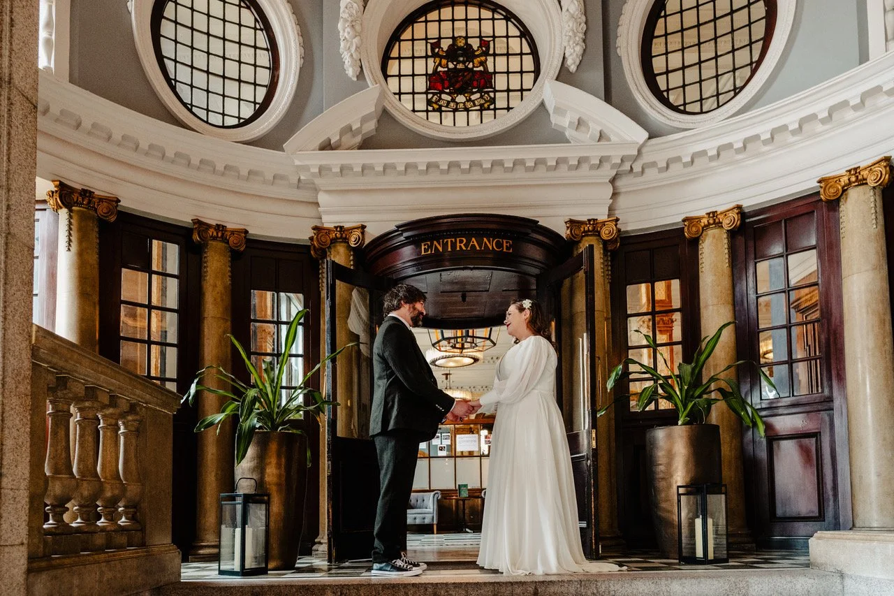 The couple stands hand in hand beneath the grand entrance of an ornate historic building, framed by tall columns, stained glass and warm wooden doors as soft light spills over their modern yet romantic outfits creating a scene that feels both timeles